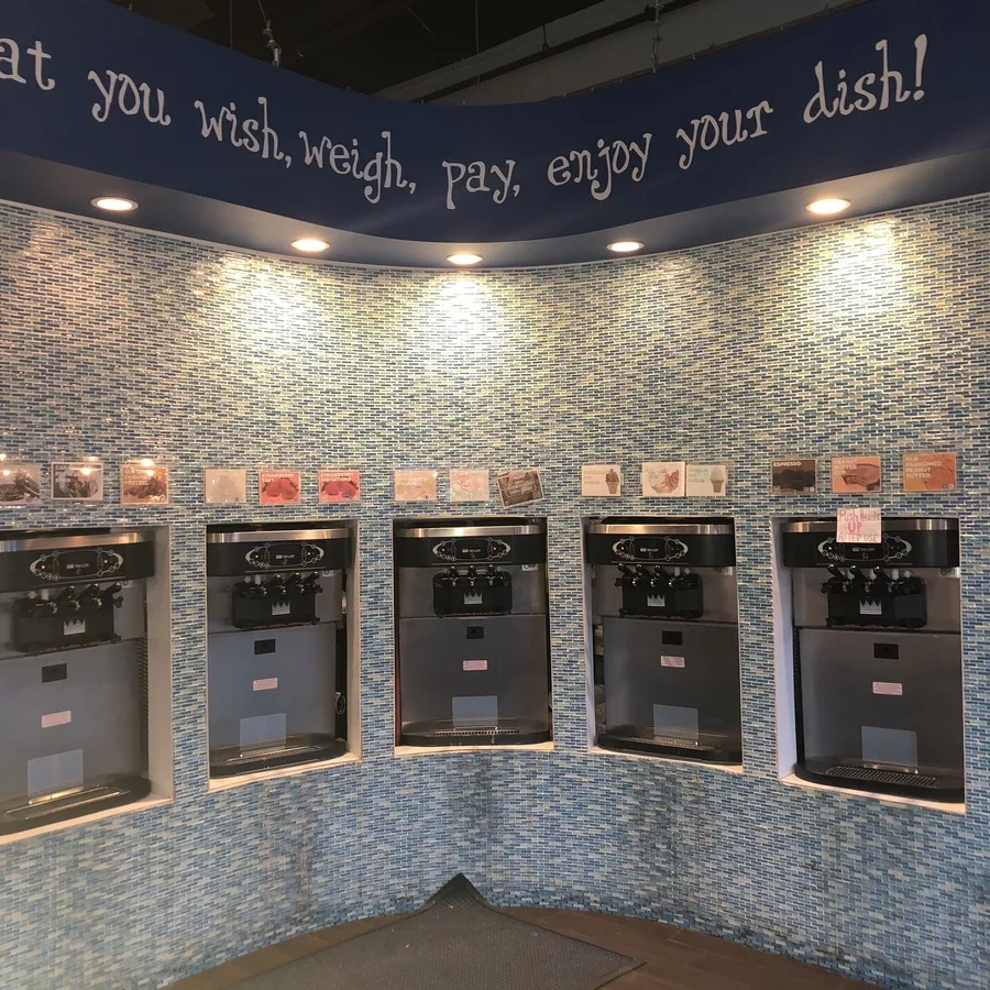 The curved tile wall of self-serve frozen yogurt machines at Sweet Waves, with the words wish, weigh, pay, enjoy your dish painted above