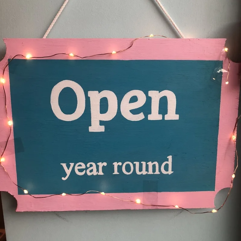 Hand-painted teal and pink sign with fairy lights that reads Open year round