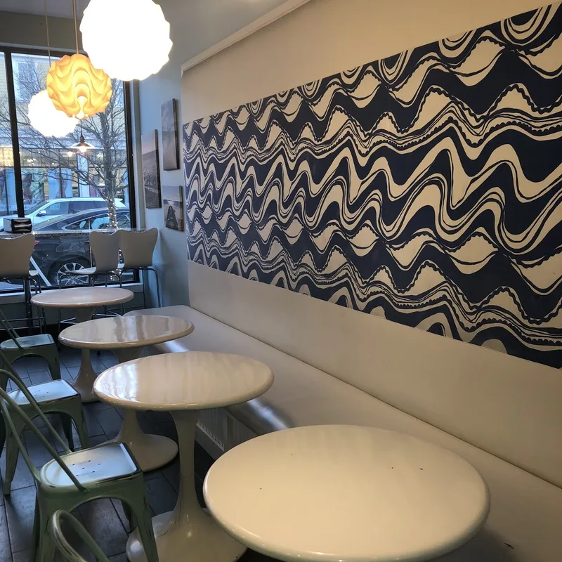 Round white tables and green metal chairs beneath a large blue and white wave mural inside Sweet Waves