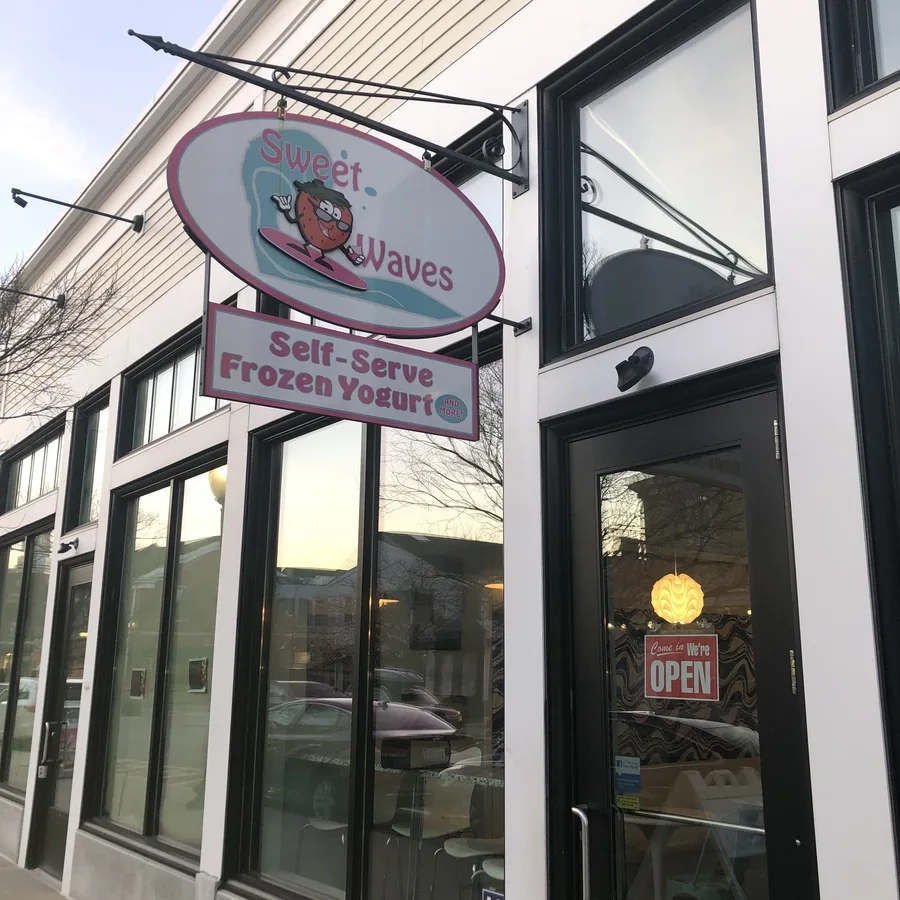 The Sweet Waves storefront with its hanging oval sign of a surfing strawberry and a Self-Serve Frozen Yogurt banner beneath